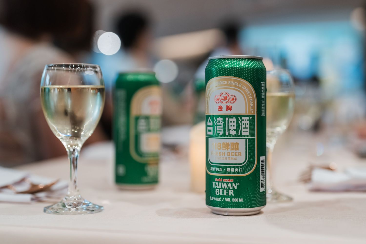 Canned beer and wine glasses arranged on a white tablecloth at a dining event.
