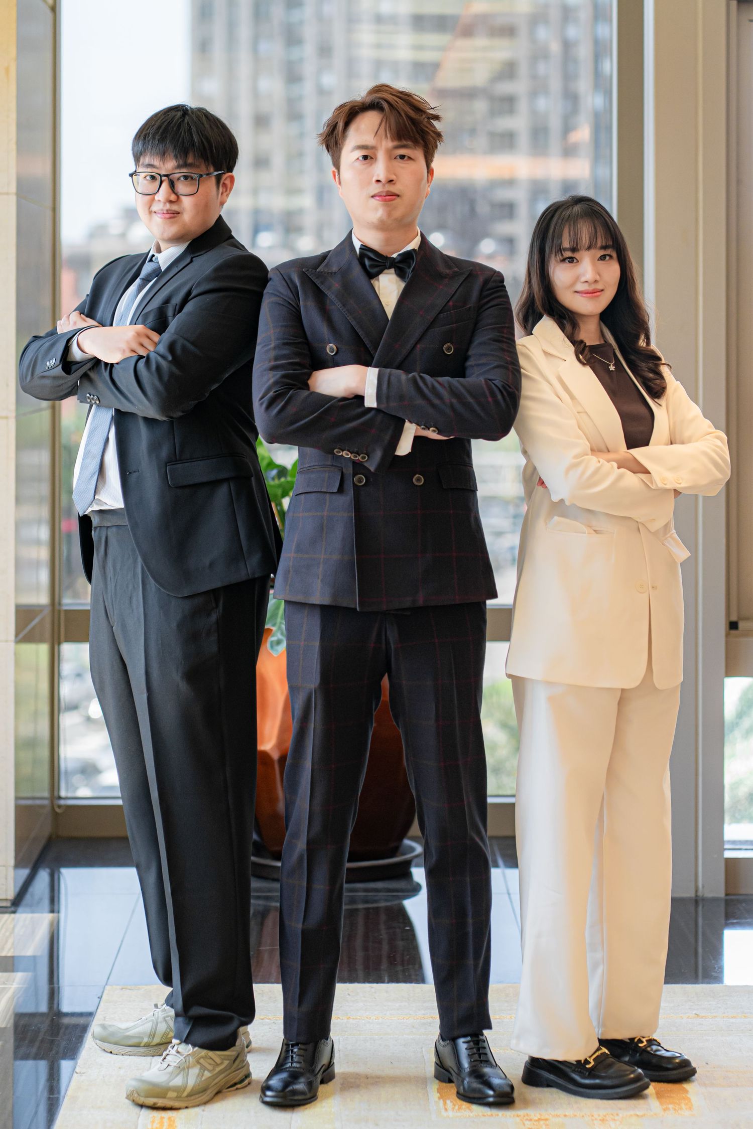 Three young Asian business professionals in formal attire standing confidently with arms crossed in a modern office.