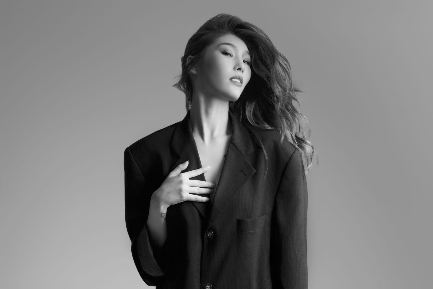 Black and white portrait of a woman in a black blazer posing gracefully with hand on chest against gray background.