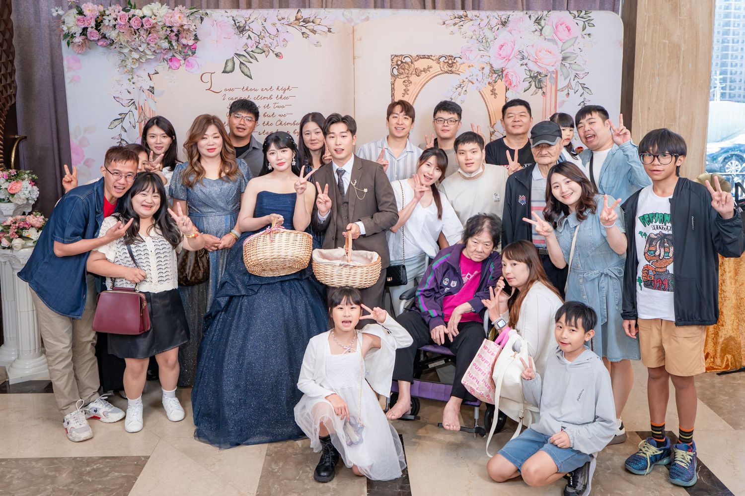 Large group of guests posing together at an elegant wedding reception with floral decorations in the background.