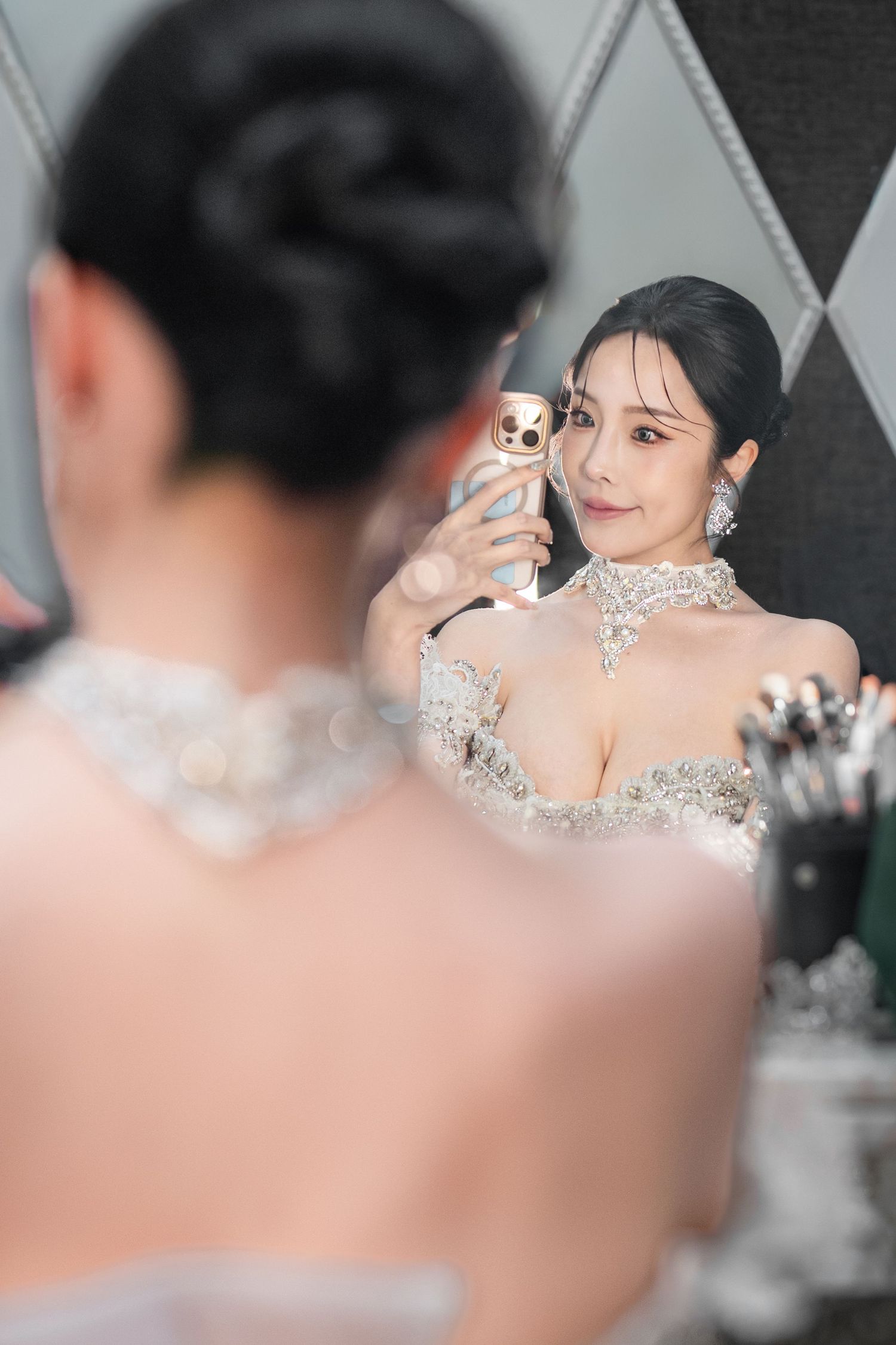 A person in a glamorous beaded gown takes a selfie in the mirror while wearing elegant jewelry and makeup.