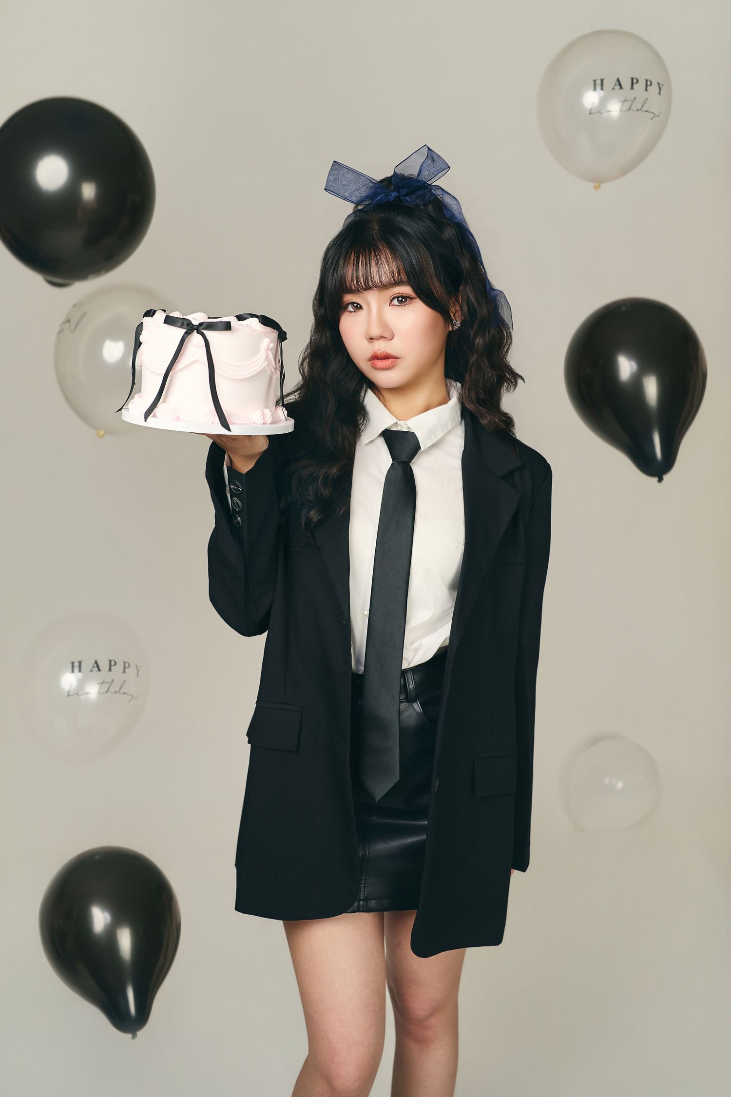 A person in a black blazer and tie holds up a white cake against a backdrop of black and white party balloons.