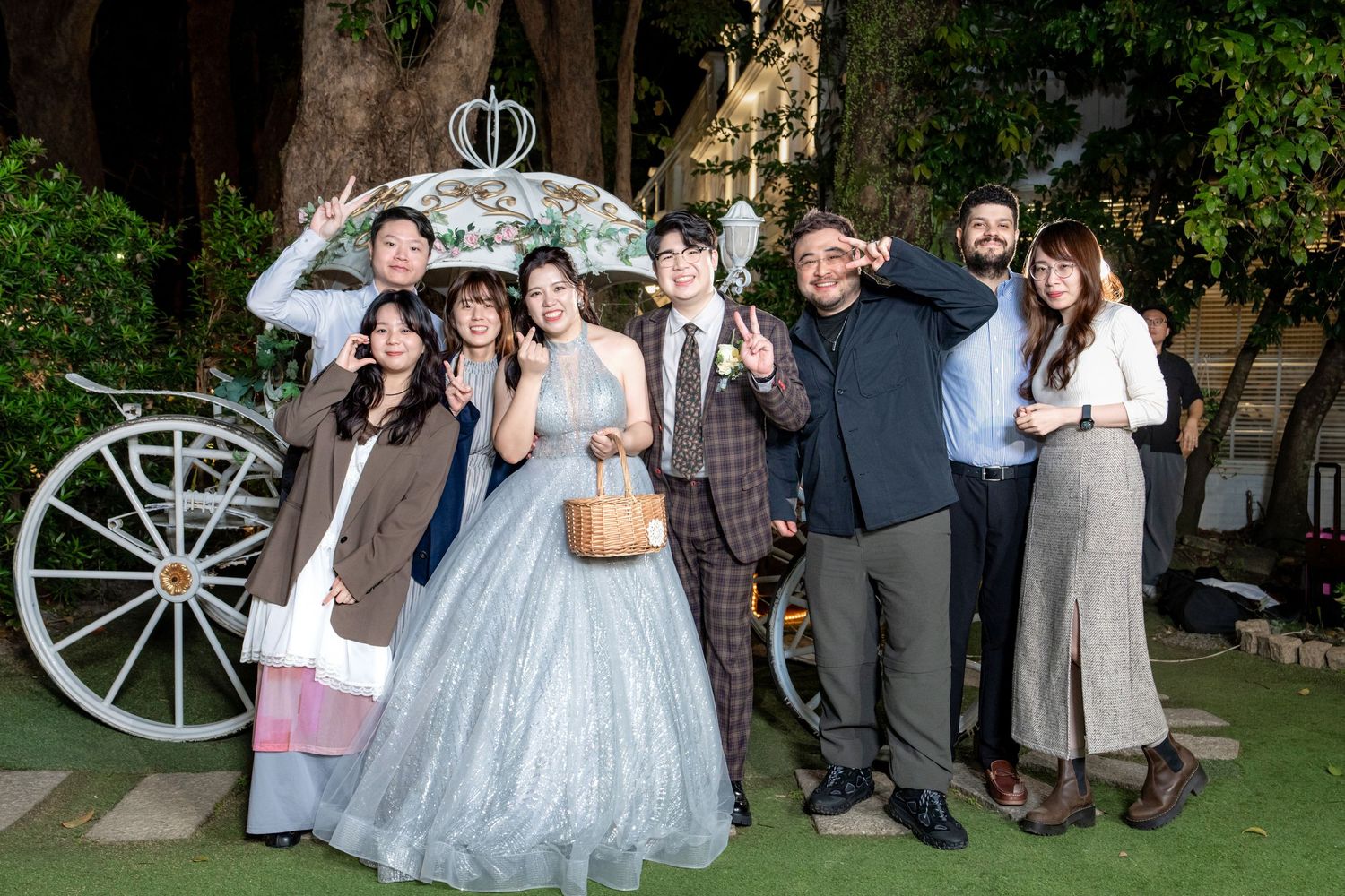 Wedding guests gather outdoors on a lawn during a celebration with a vintage carriage wheel visible in the background.