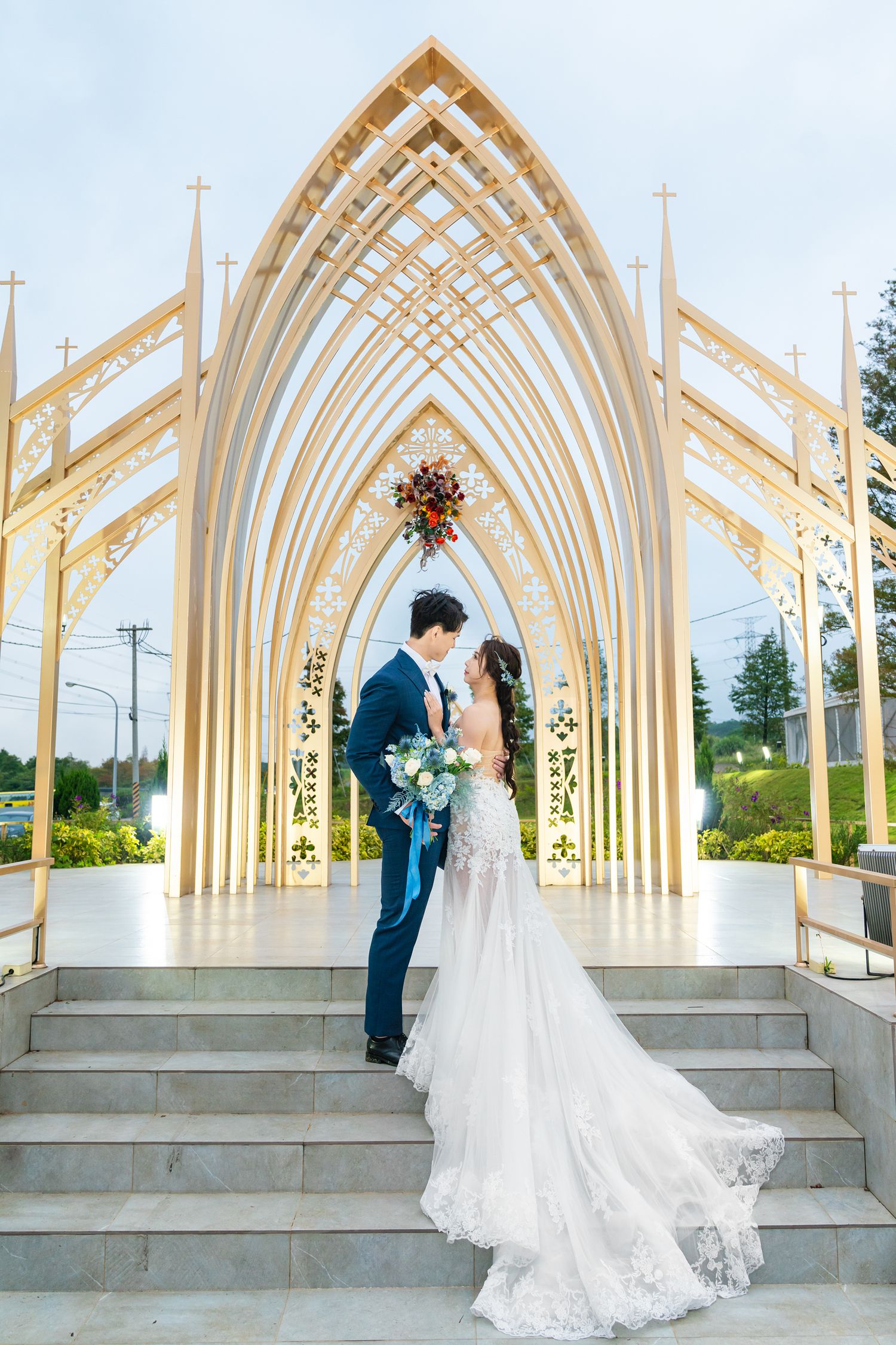 A couple embraces under a stunning modern white cathedral arch with decorative floral elements at an outdoor wedding venue.