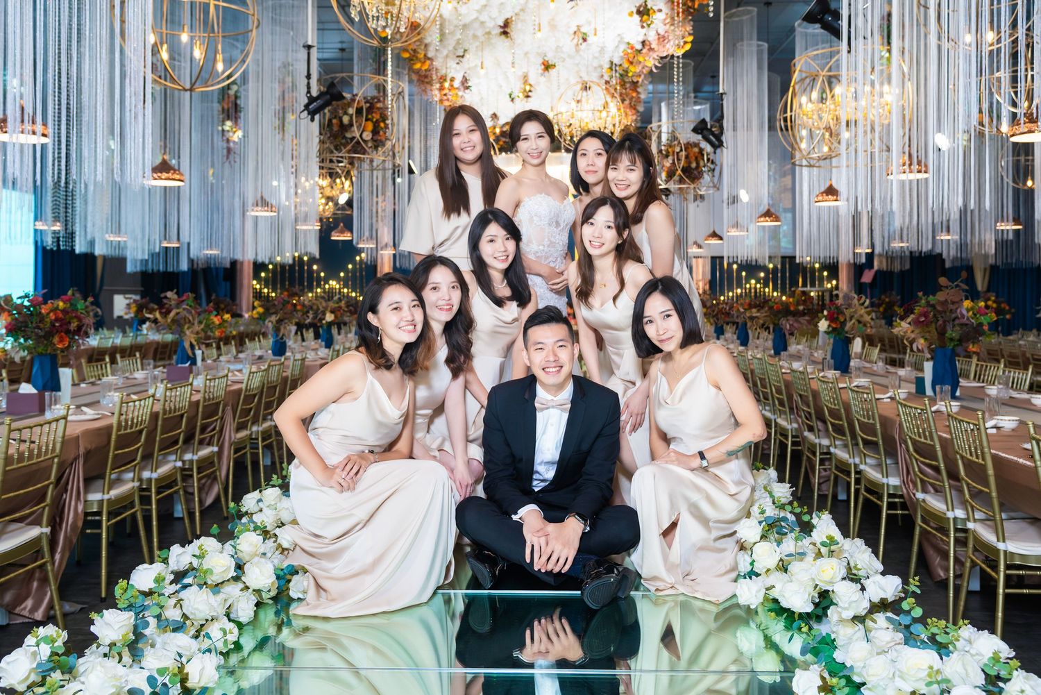 Elegant wedding party group posed in luxurious banquet hall with crystal chandeliers and floral decor.