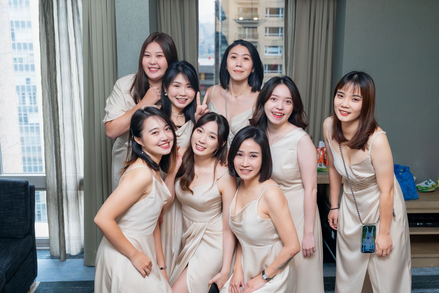 A group of bridesmaids pose together wearing matching champagne-colored silk dresses in a bright hotel room.