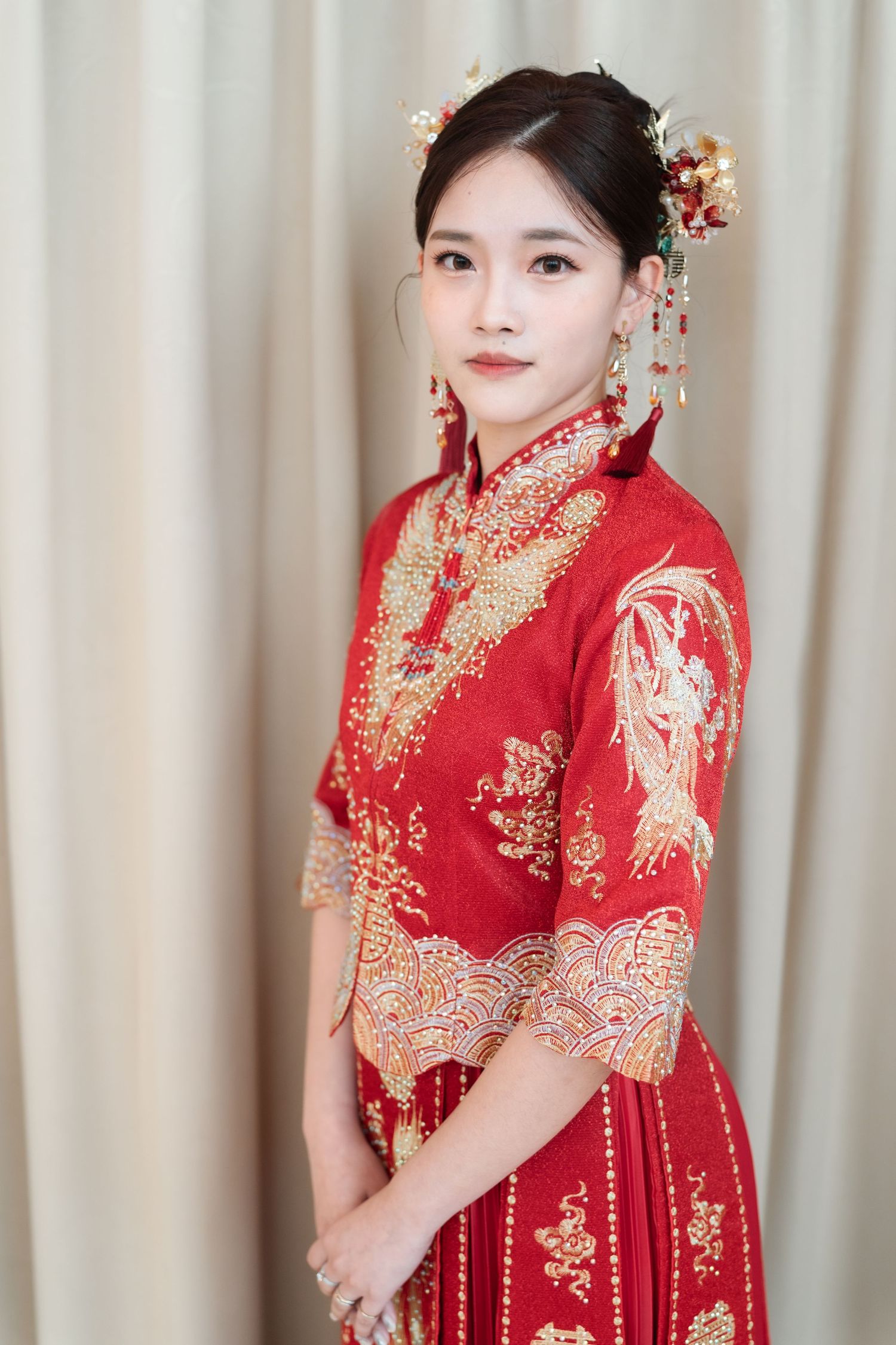 A person wearing a traditional red Chinese wedding dress with gold embroidery and floral hair accessories poses elegantly.