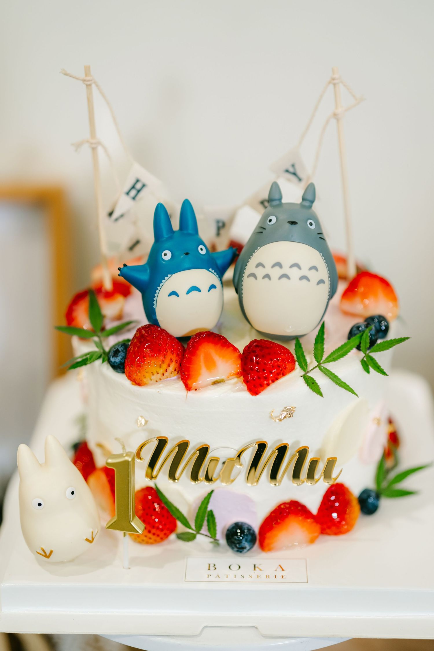 A white frosted cake decorated with Totoro characters, fresh strawberries, and blueberries on a cake stand.