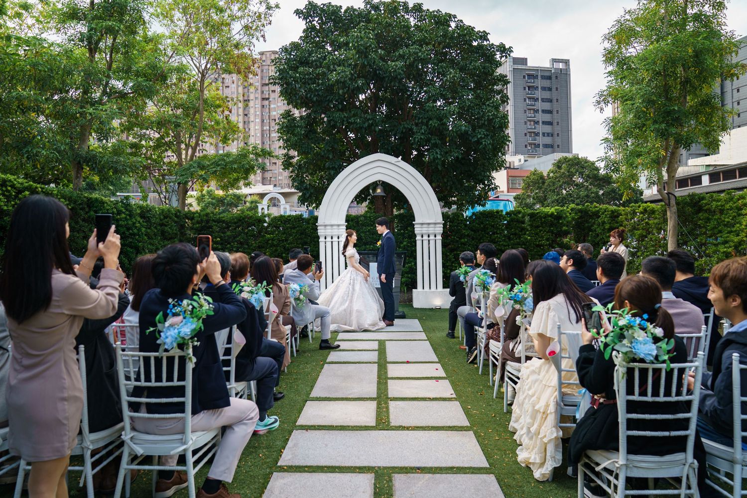 Wedding guests seated in white chairs watch an outdoor ceremony in a lush garden setting with tall trees and columns.