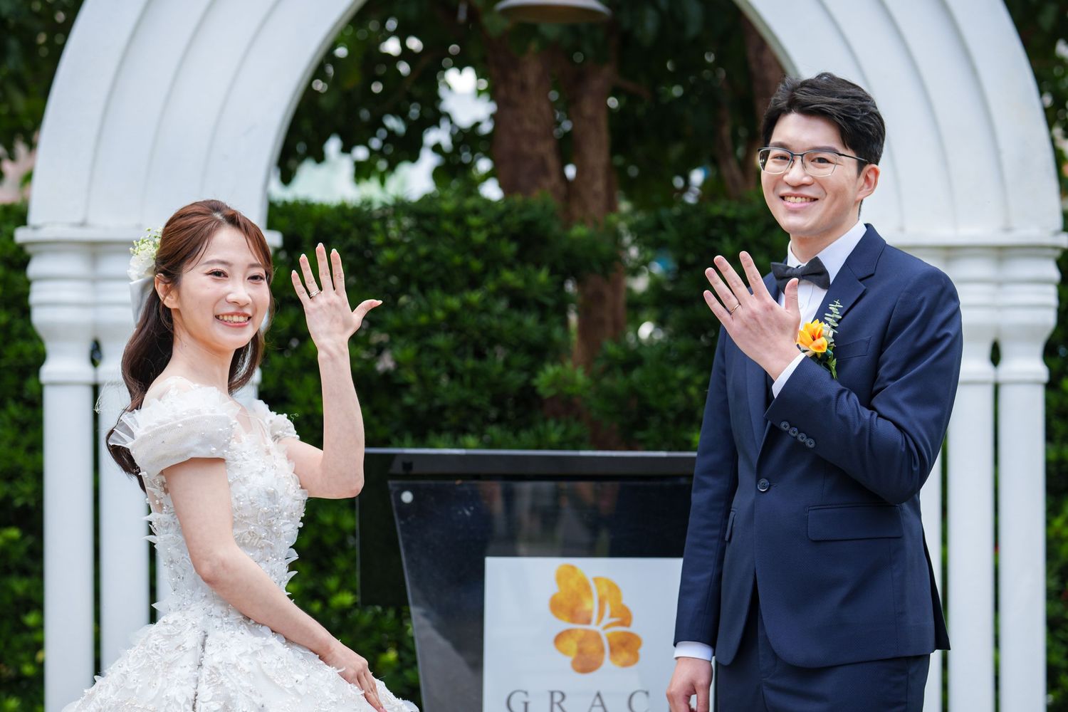 A happy bride and groom wave to guests during their outdoor wedding ceremony near white pillars.