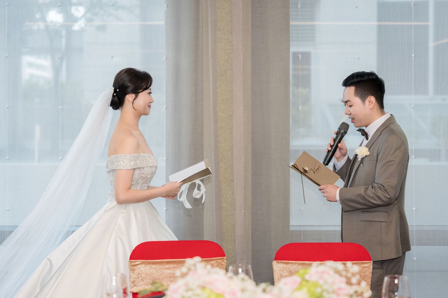 A couple in wedding attire reads from papers during their ceremony in a bright indoor venue.