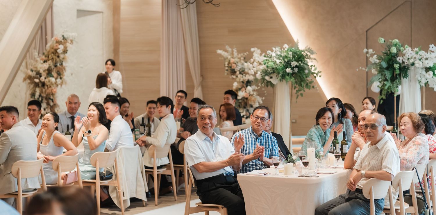 Wedding reception guests seated at round tables with elegant white floral centerpieces in a light-filled venue.