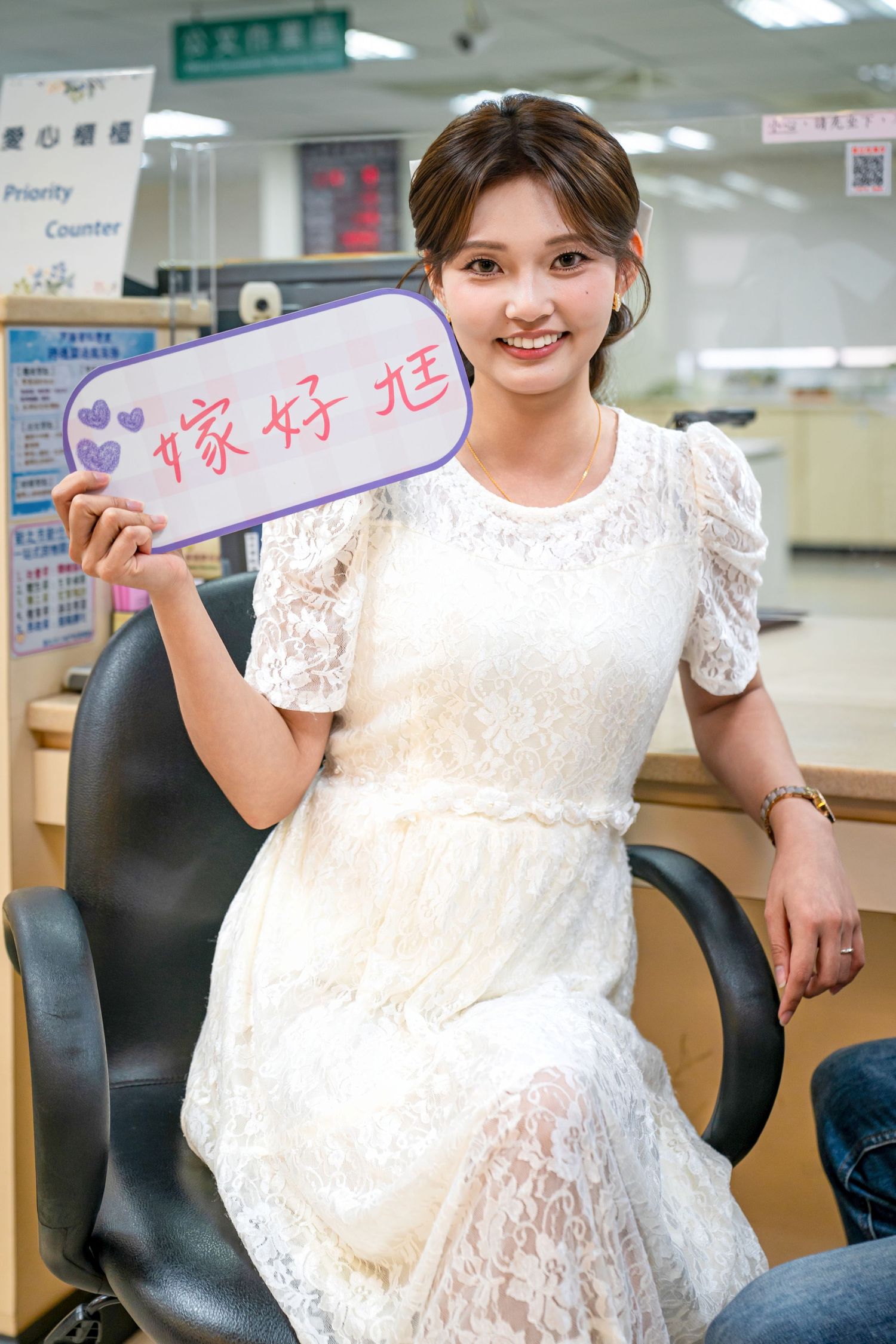 Person in white lace dress sitting in office chair holding up sign with Chinese characters.