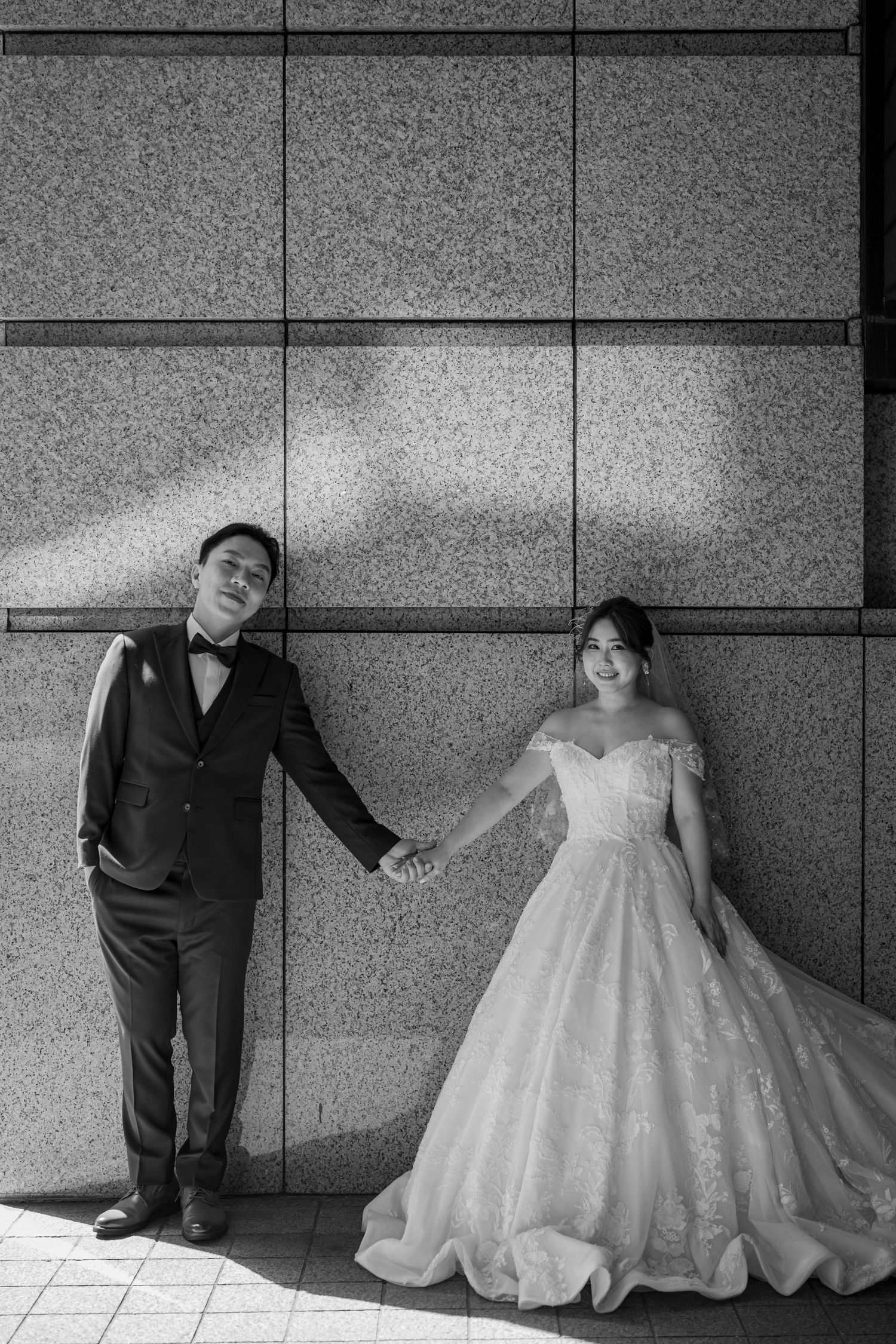 A black and white photo series showing a couple in formal wedding attire posing playfully against a modern wall.