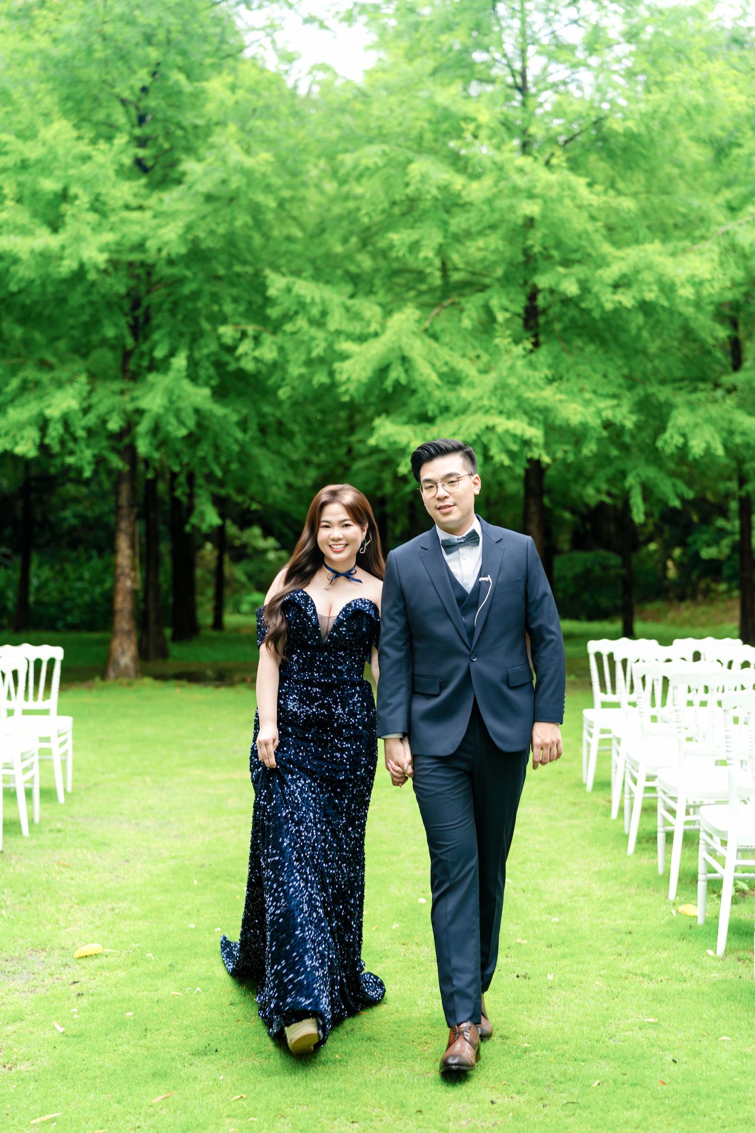 A couple in formal attire walks along a garden path with rows of white chairs set up for an outdoor wedding ceremony.