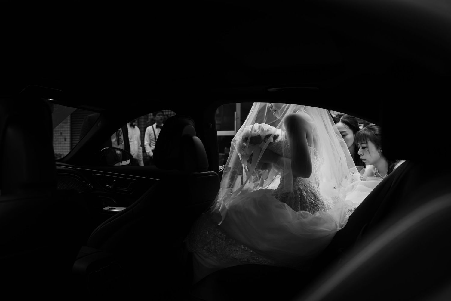 Artistic black and white photo of bride in wedding dress seen through car window reflection.