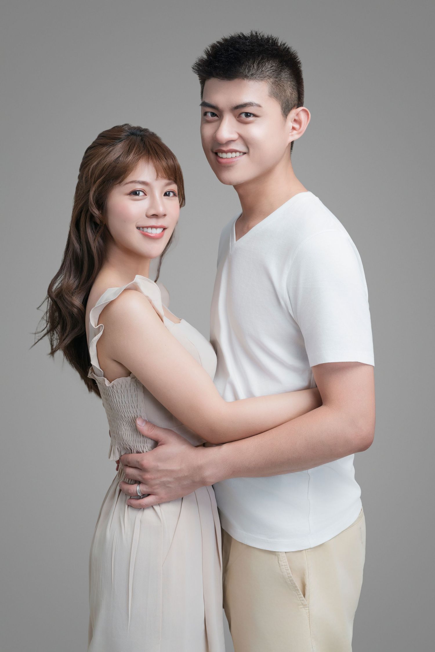 A young couple in white and beige clothing embrace while posing for a romantic studio portrait against a gray background.