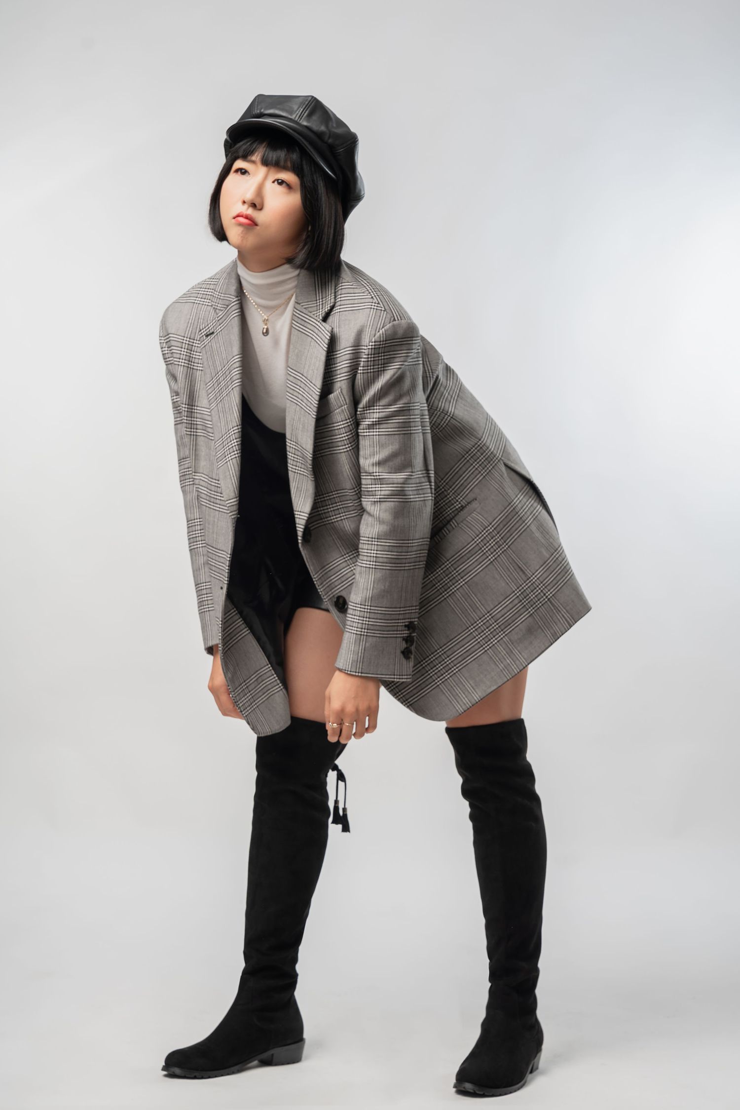 Model wearing gray plaid blazer, turtleneck, black shorts and thigh-high boots with black cap in fashion photoshoot.