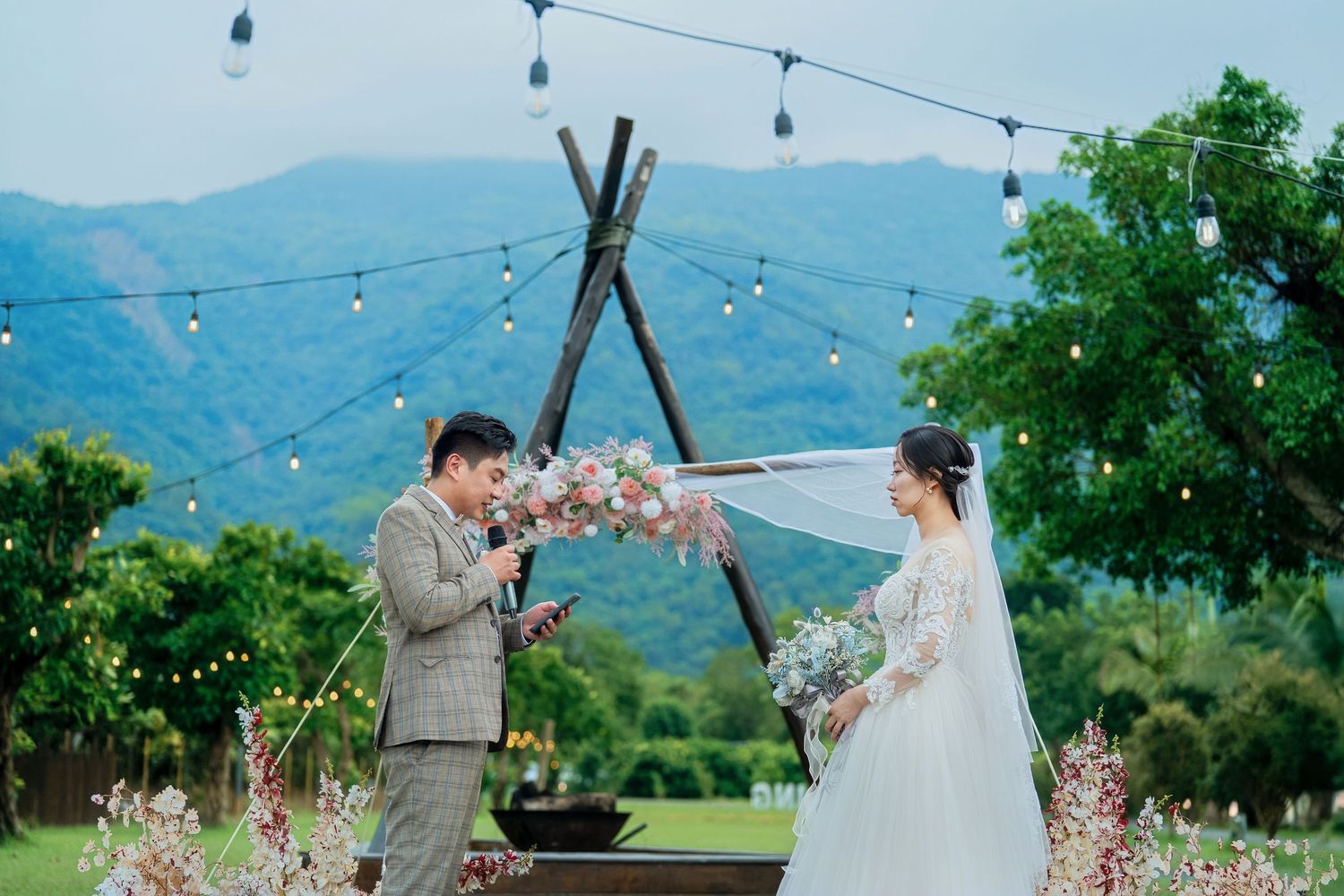 Romantic outdoor wedding ceremony under string lights with mountain views and blooming flowers in the background.
