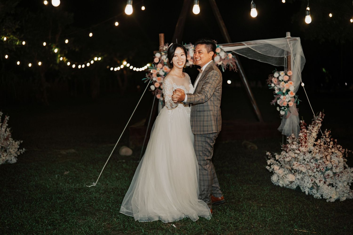 Outdoor wedding celebration at night with string lights glowing in the background and floral decorations.