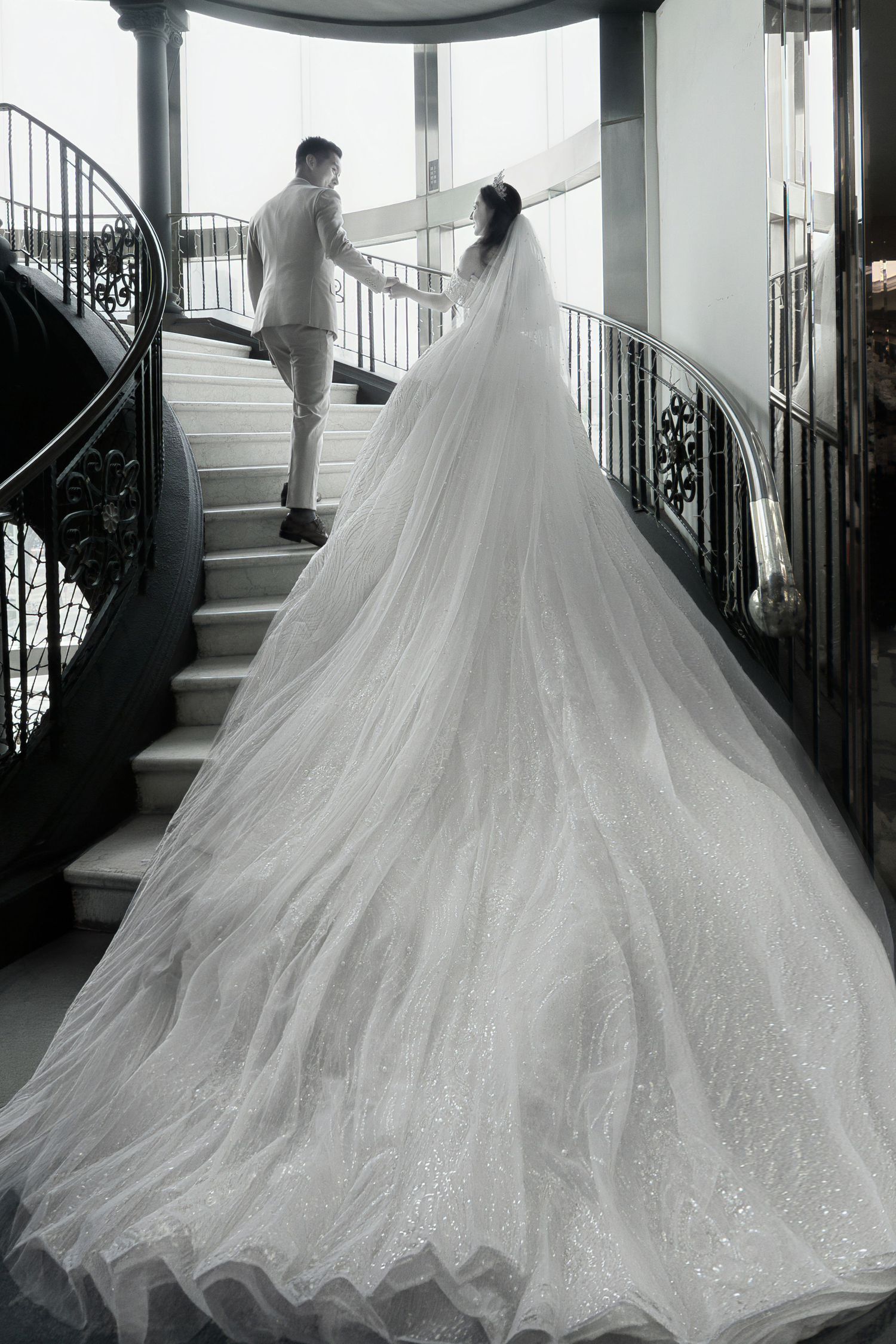 Bride in flowing white gown with long veil ascending grand staircase with groom, elegant black and white wedding photo.