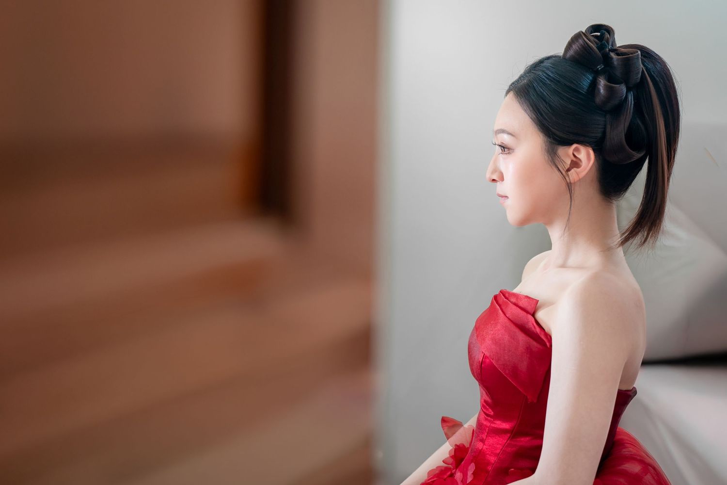 Young woman in elegant red strapless dress with updo hairstyle sitting gracefully near staircase in profile view.