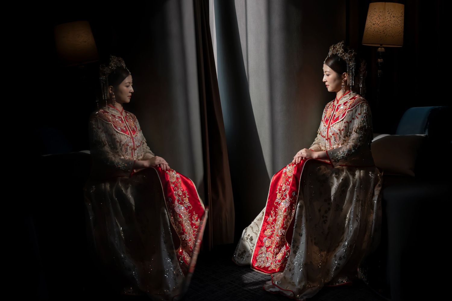 A bride in a red and silver traditional Asian gown sits reflected in a mirror by draped curtains in dim light.