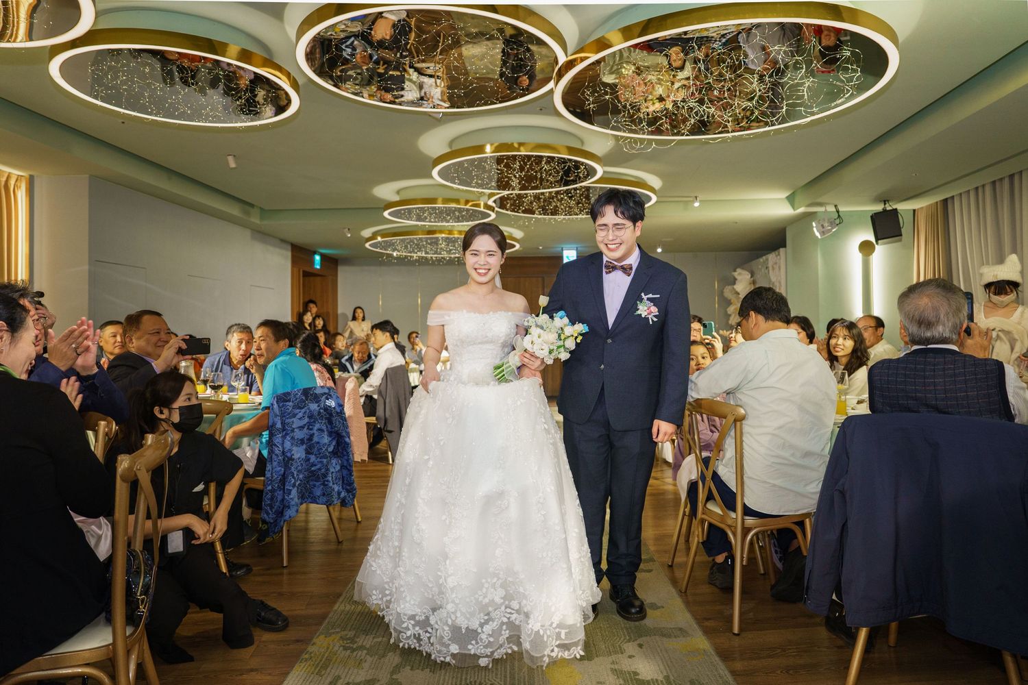 Wedding reception guests dancing and celebrating in an elegant ballroom with circular ceiling details.