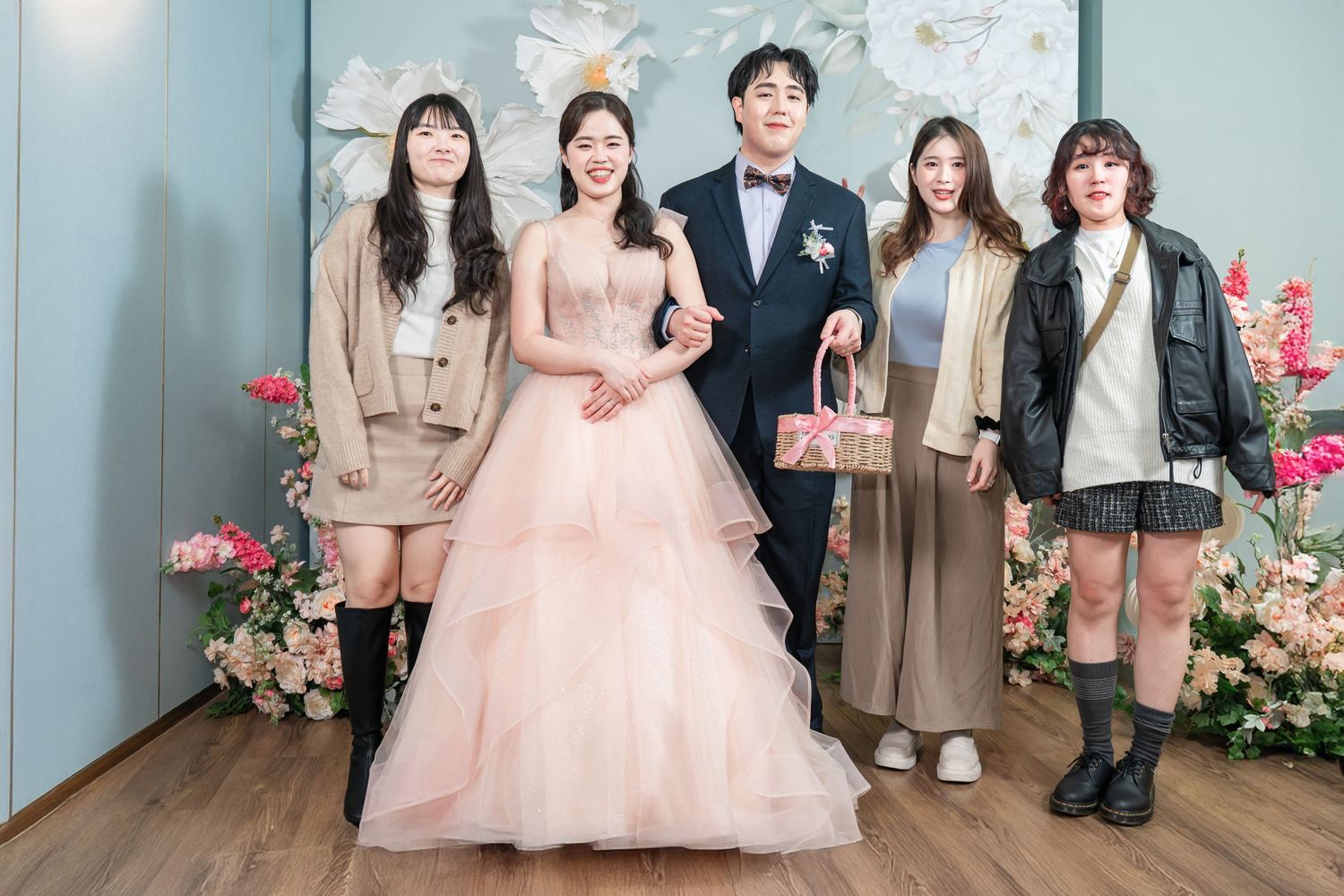 A group of people pose together in formal attire against a floral backdrop in an indoor setting.