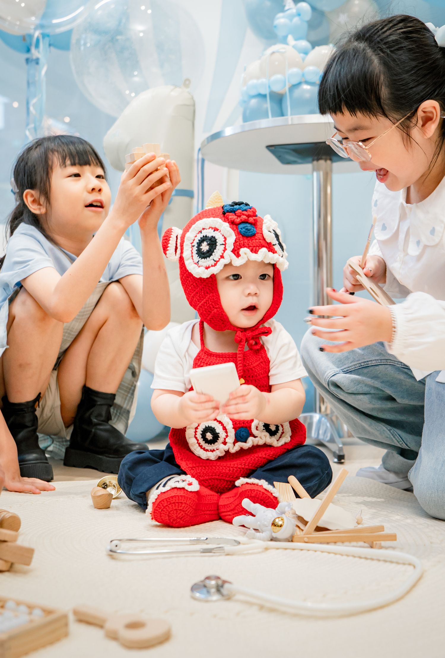 A child in a red outfit plays with wooden toys while lying on the floor in a bright indoor setting.