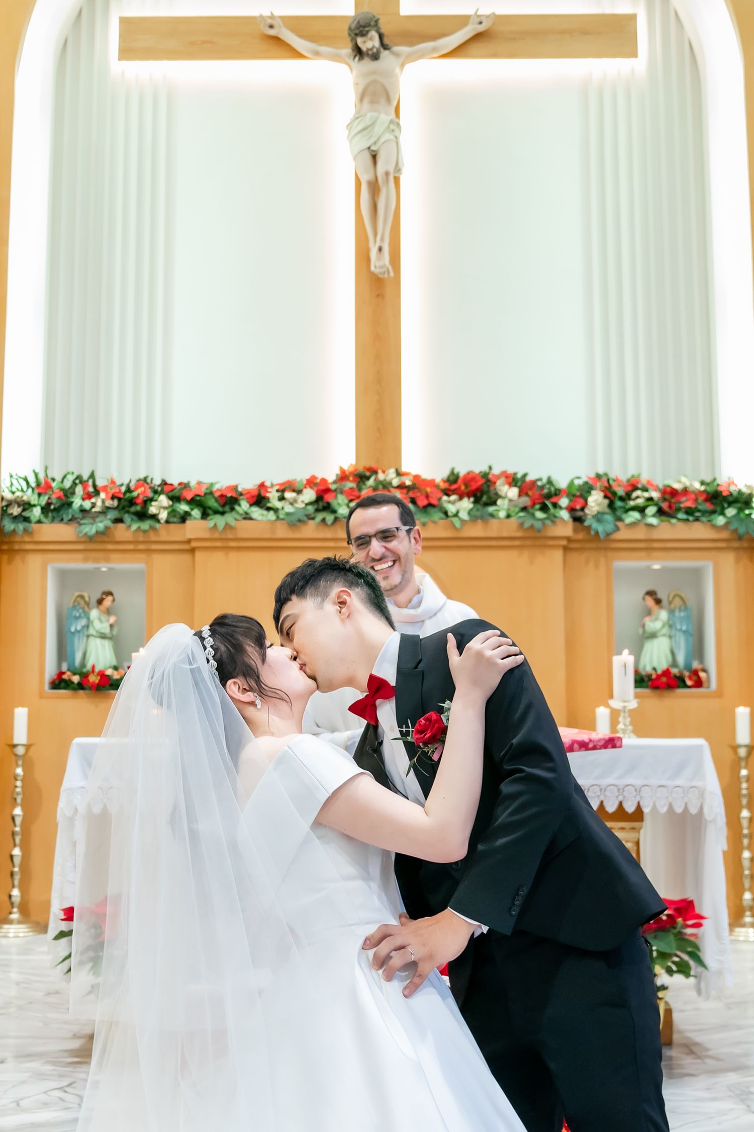 A wedding ceremony takes place in a Catholic church adorned with festive red and green floral decorations.
