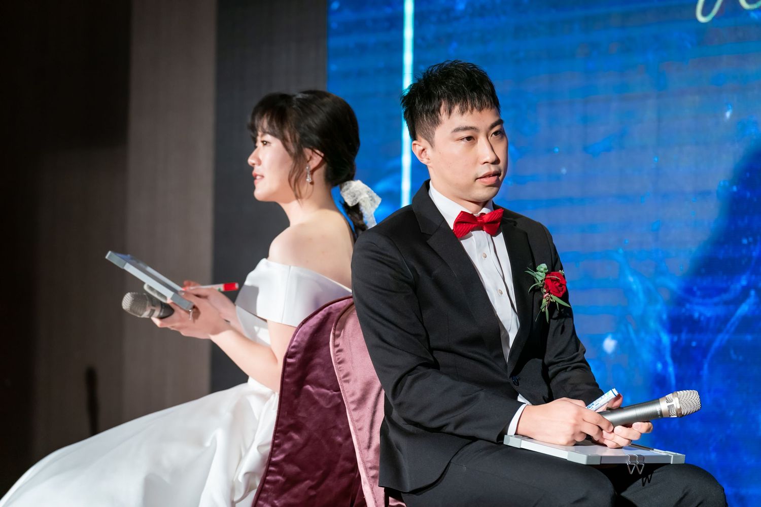 A bride and groom sit back-to-back at their wedding reception holding notebooks against a blue backdrop.