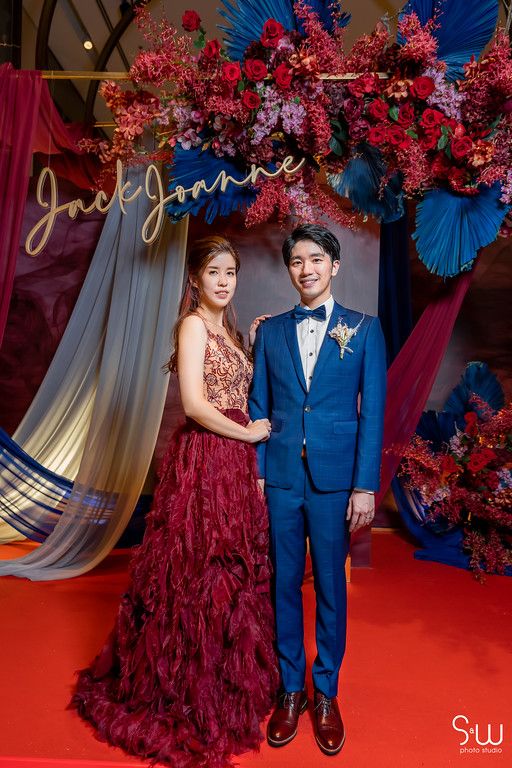 Formal portrait features a burgundy gown and blue suit against dramatic red and blue floral backdrop.