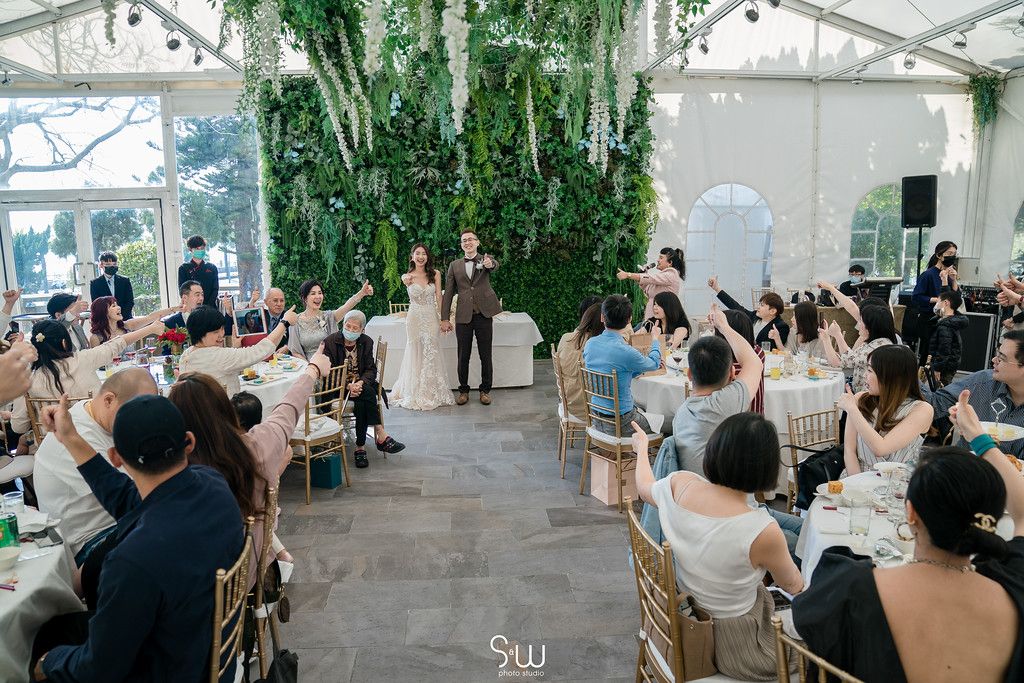 Guests raise their hands during a wedding ceremony in a greenhouse-style venue with hanging greenery.