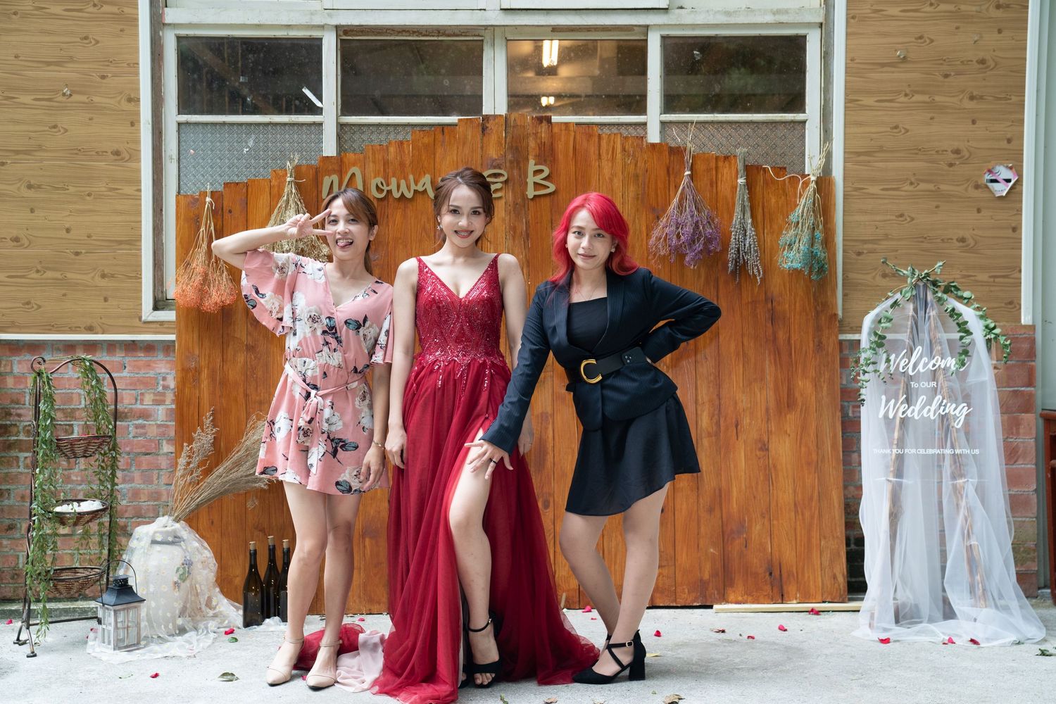 Models in floral, red, and black outfits pose playfully against a wooden wall with dried flowers.