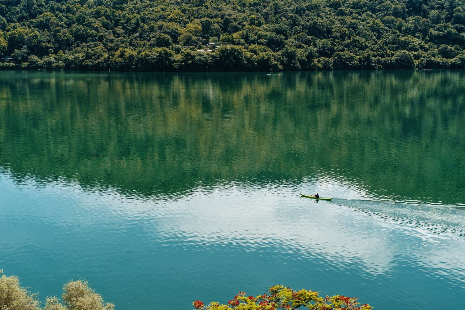 Lone kayaker gliding across a serene green lake surrounded by forested hills.