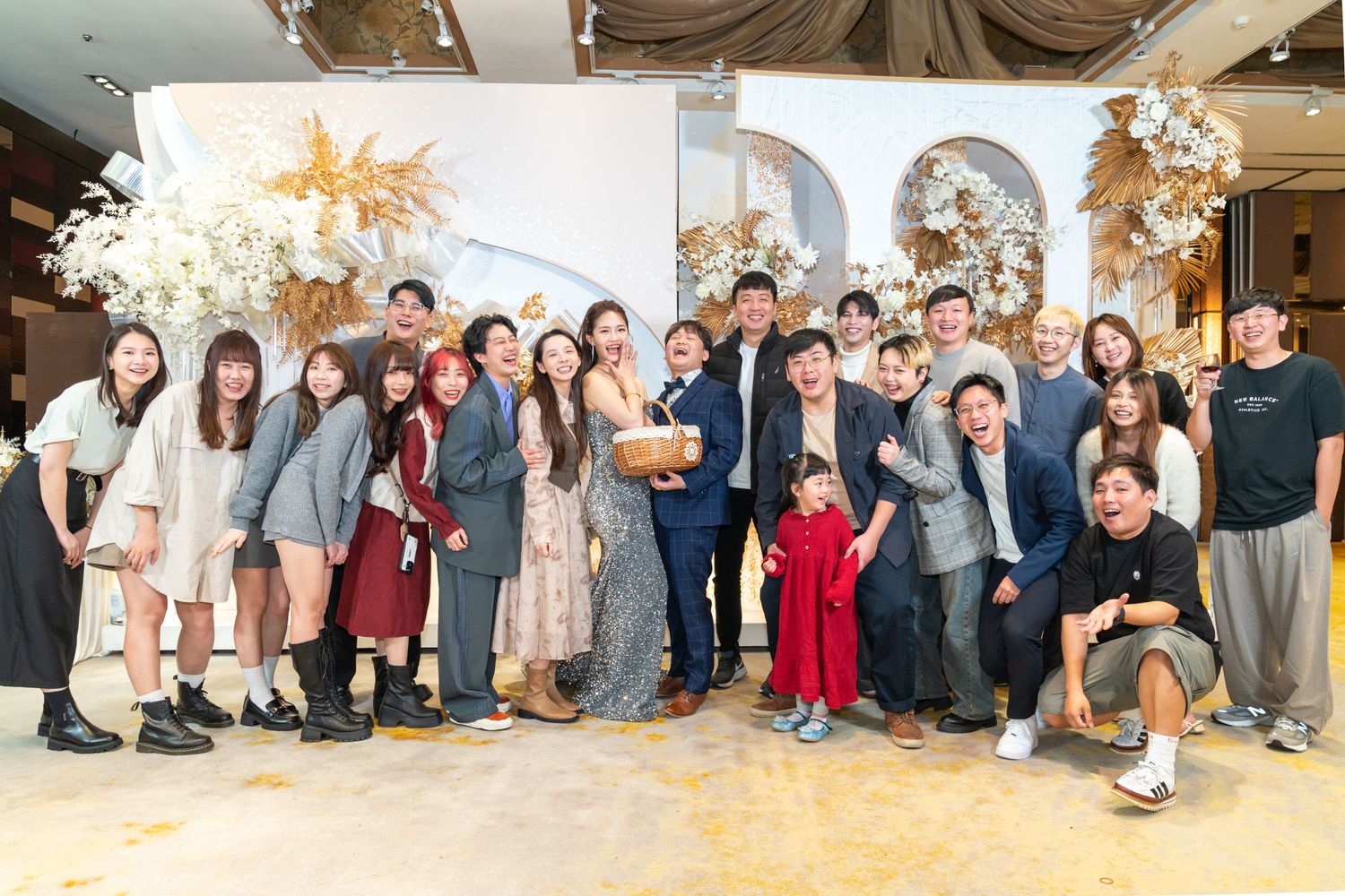 A large group celebrates together in an elegant venue decorated with white floral arrangements and golden details.