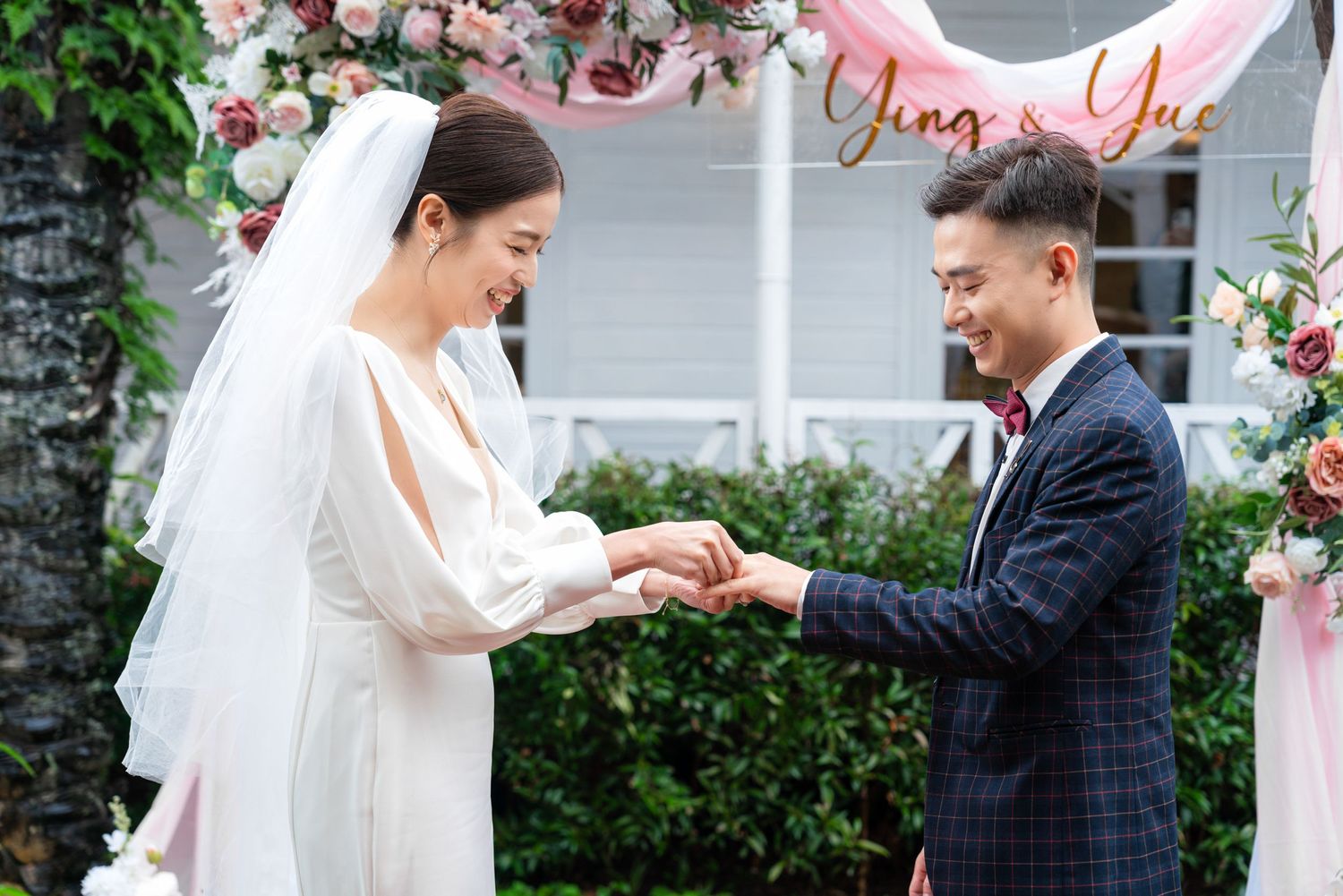 A joyful outdoor wedding ceremony with a couple exchanging rings under floral decorations and greenery.