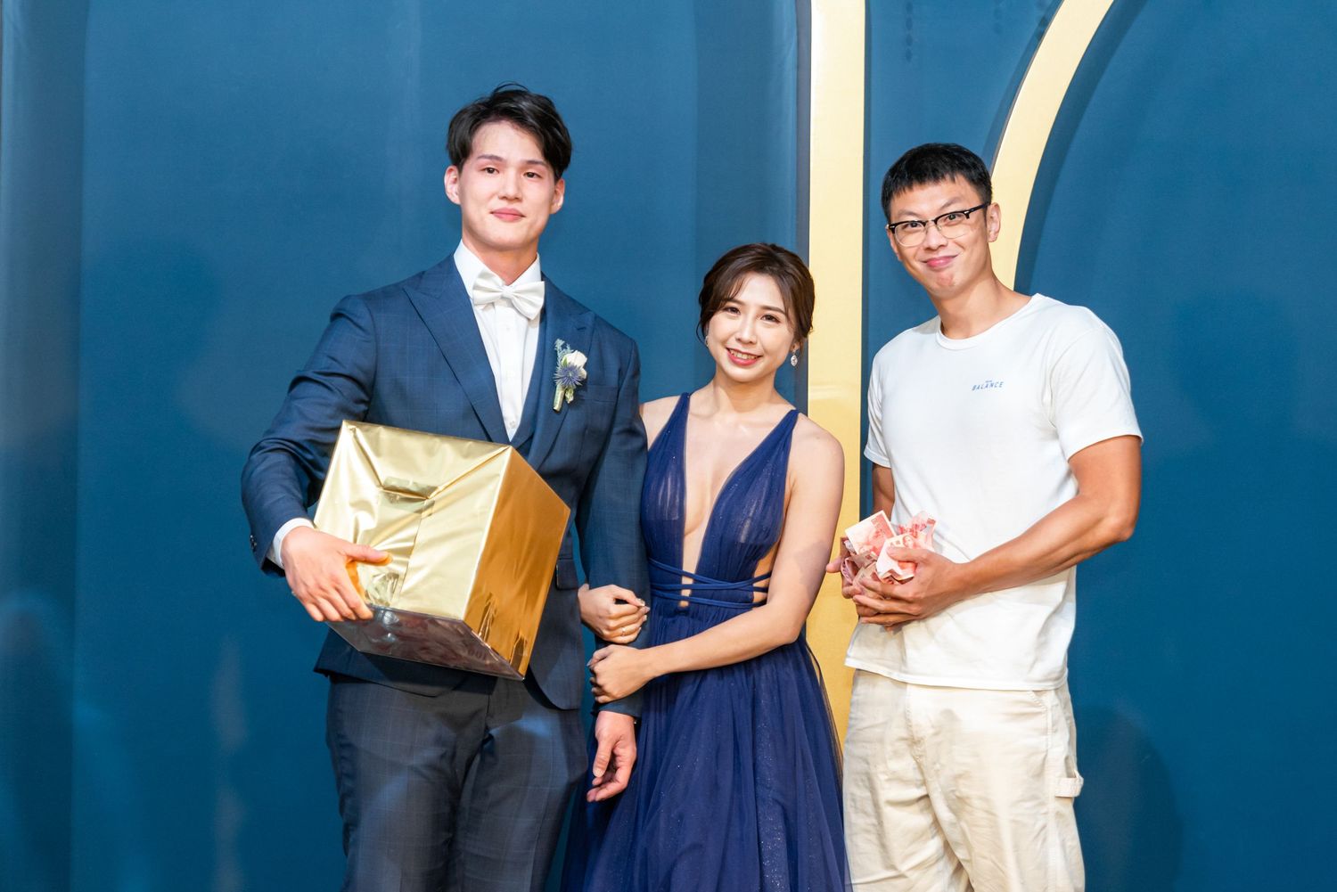 Three people stand against a blue wall backdrop, with one person in a suit, one in a navy dress, and one in white attire.