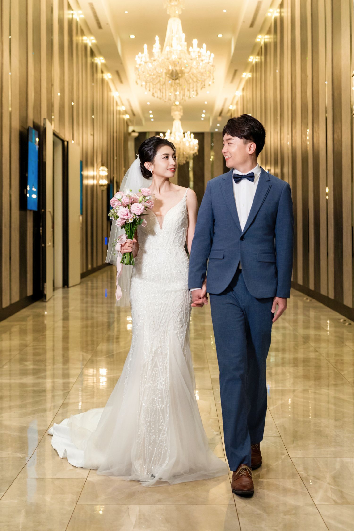 A couple in wedding attire walks down an elegant hallway with chandeliers and marble floors.