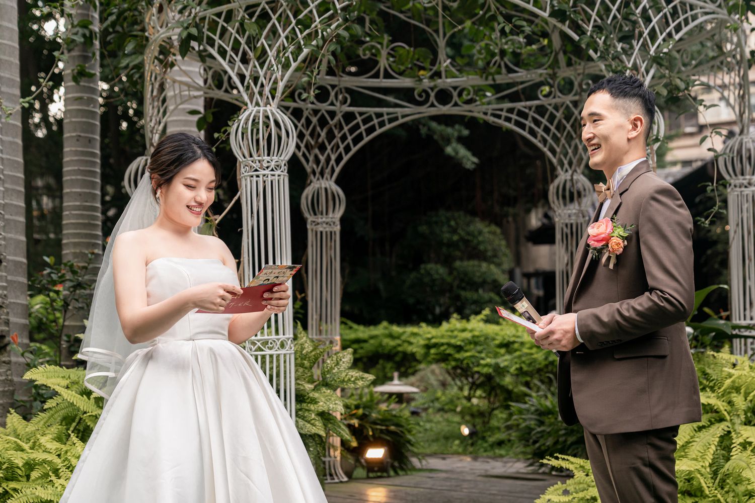 A couple exchanges vows during an outdoor wedding ceremony beneath an ornate garden arch surrounded by greenery.
