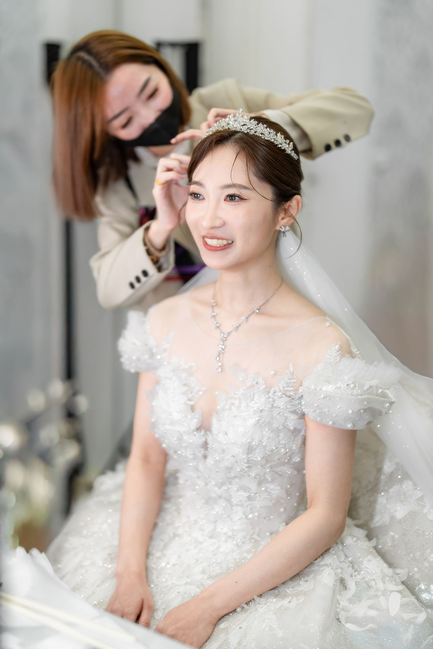 A bride wearing a white lace wedding dress and tiara gets her hair styled while smiling at the camera.