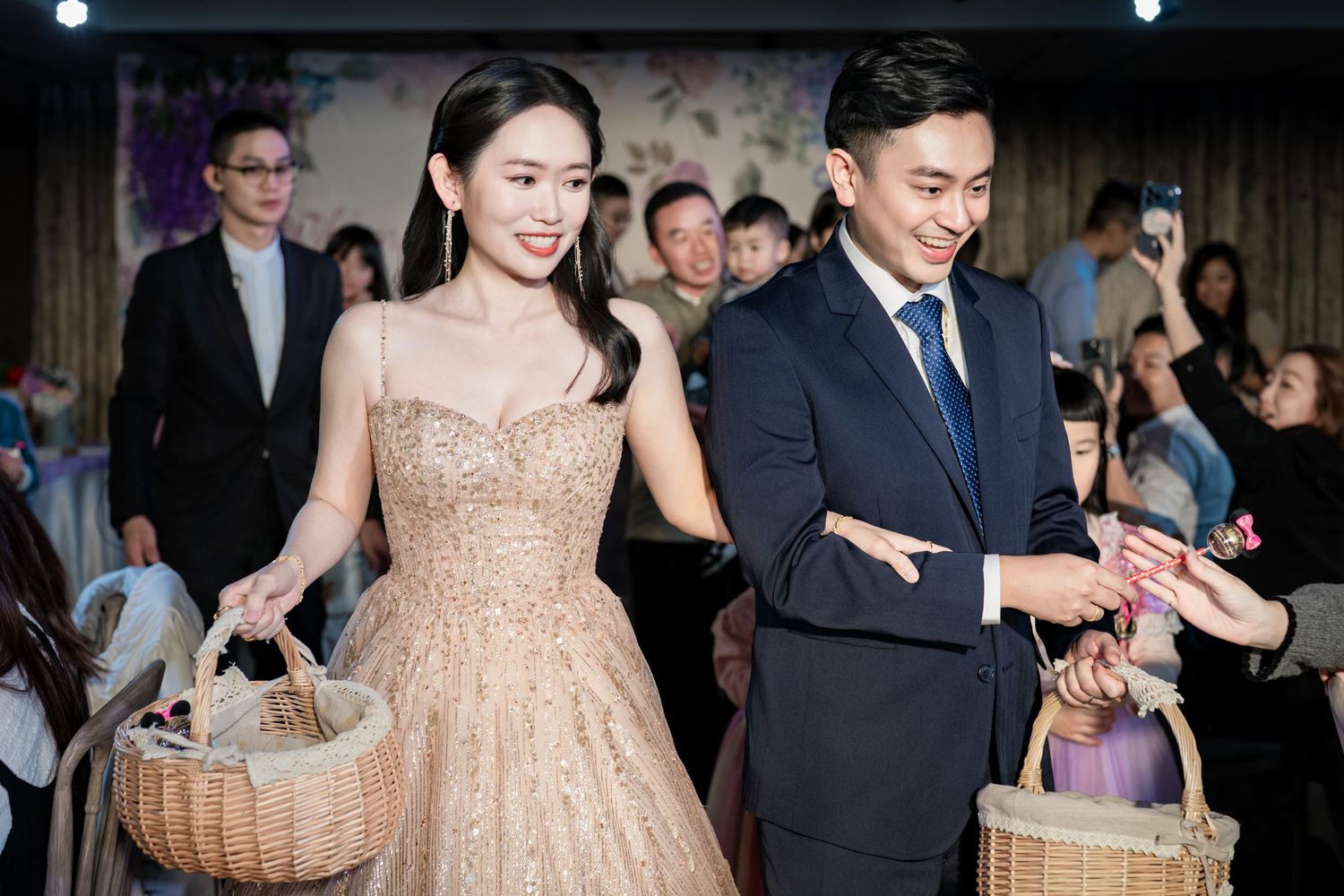 Couple in formal attire walking together at an event, carrying decorative baskets through a crowd.