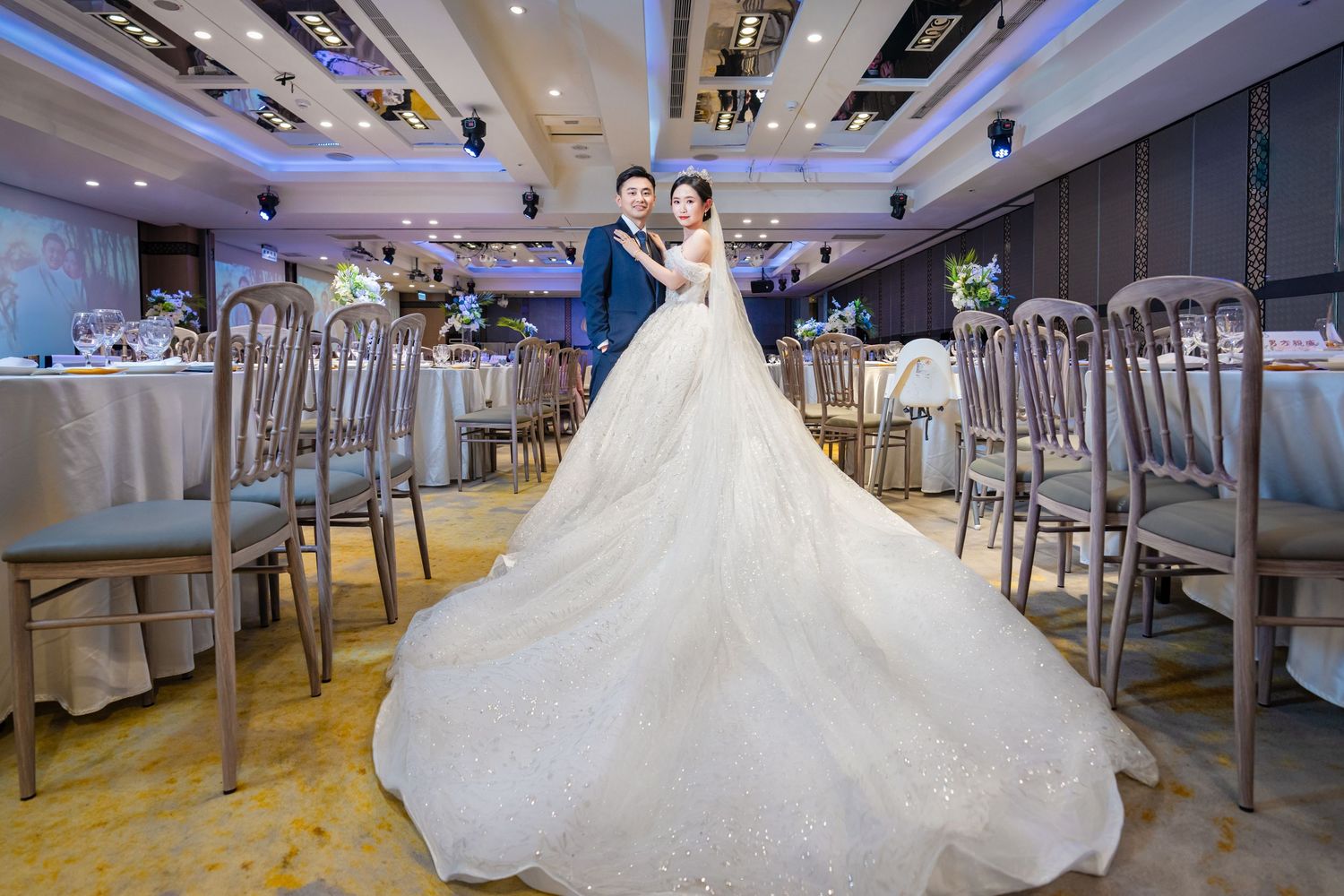 A wedding reception venue with elegant decor featuring a long row of white-clothed tables and chairs under modern lighting.