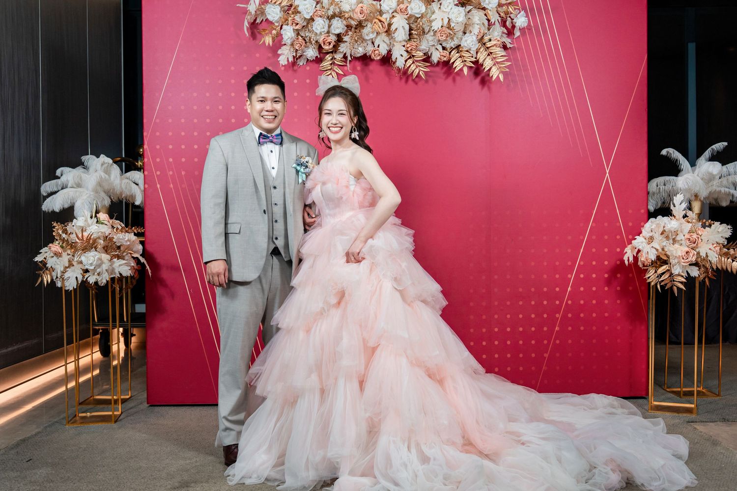 A couple poses in front of a pink backdrop with elegant floral decorations at a wedding celebration.