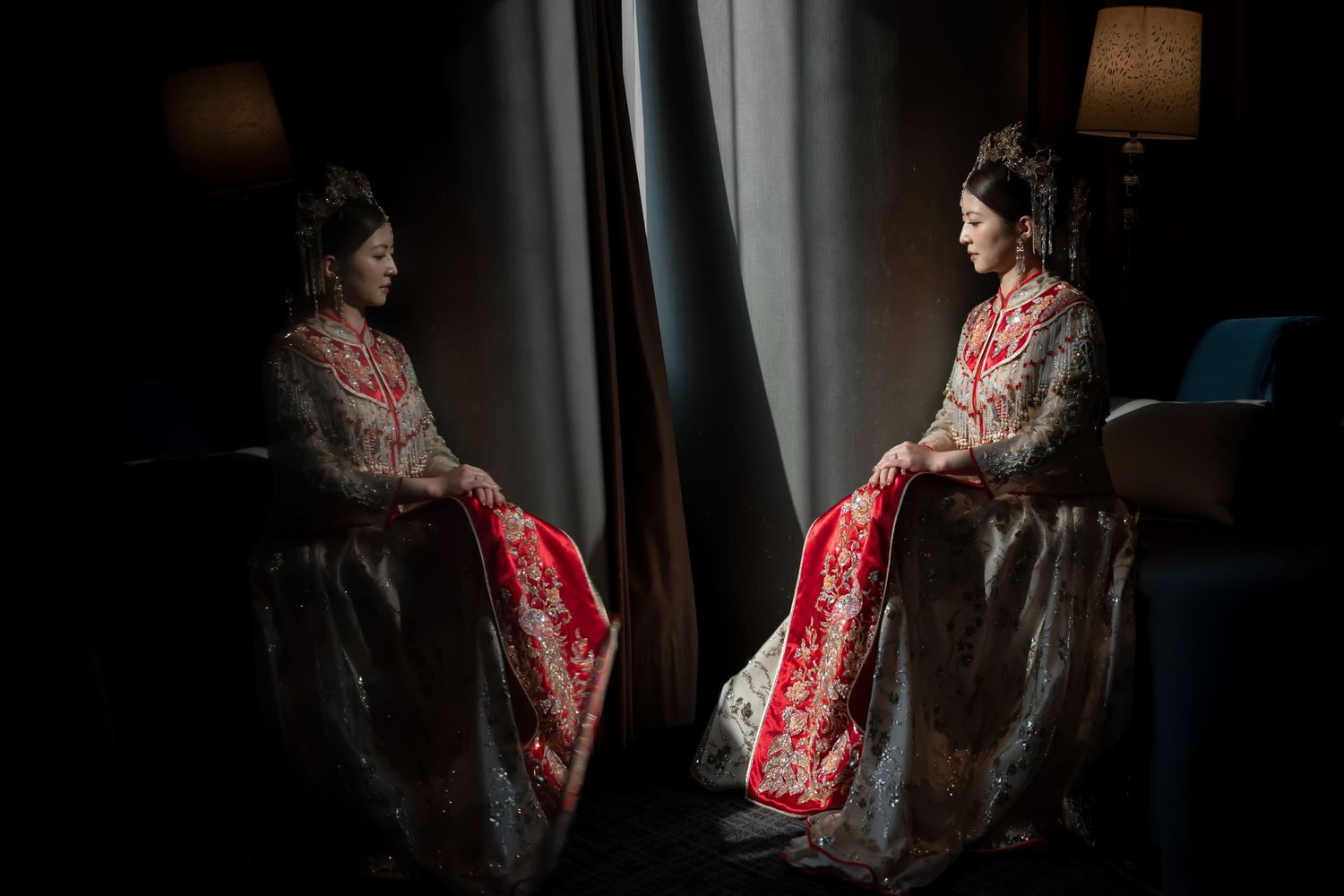 Bride in an ornate red and gold embroidered traditional gown sits beside a window, mirrored in a dramatic low-light reflection.
