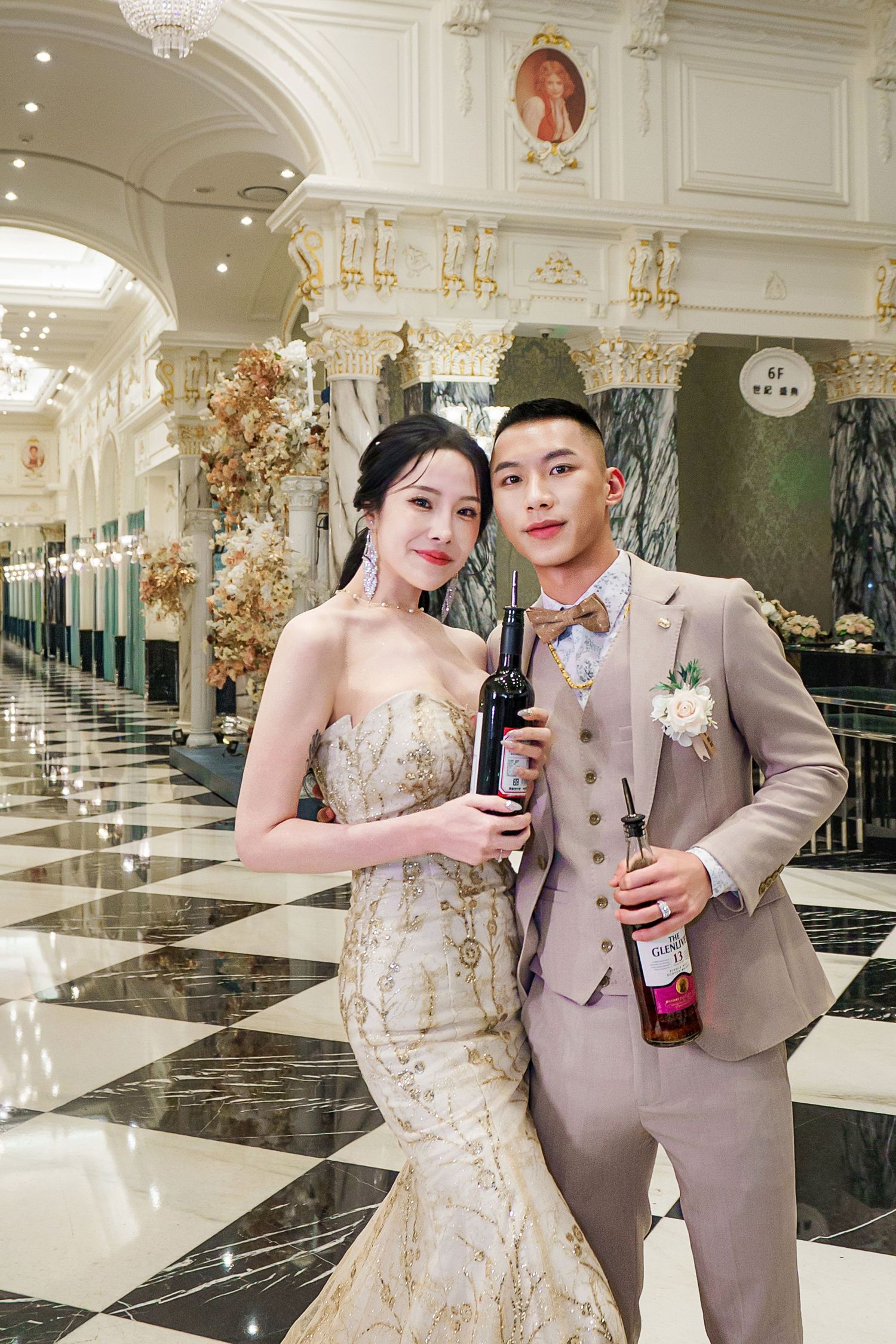 Couple in formal attire poses together in an elegant marble ballroom with ornate chandeliers and gold accents.