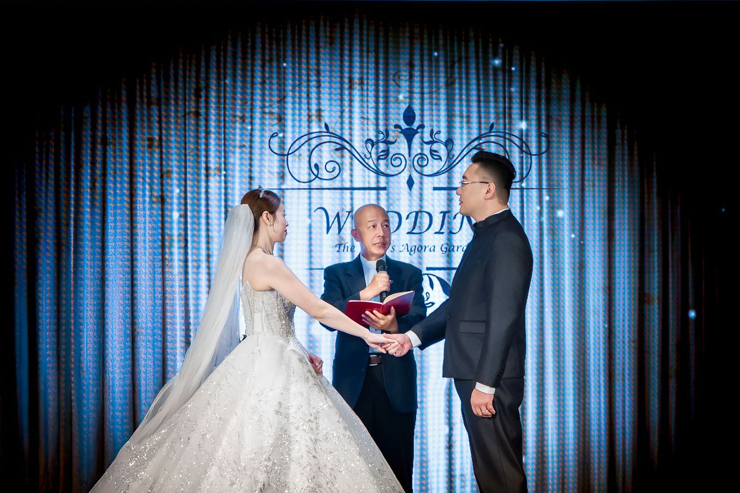 A couple in wedding attire dance together in front of a blue-lit curtain backdrop during their reception celebration.