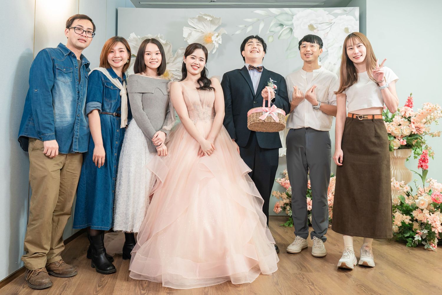 A wedding party poses together in a light-filled venue with pink and white floral decorations.