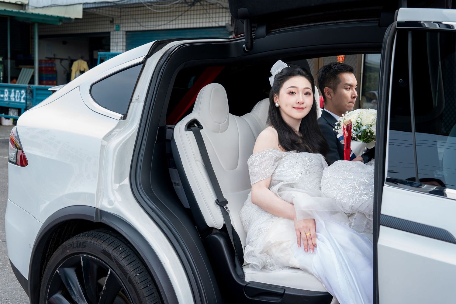 Bride in white wedding gown sitting in white Tesla Model X falcon-wing door with groom standing behind her.